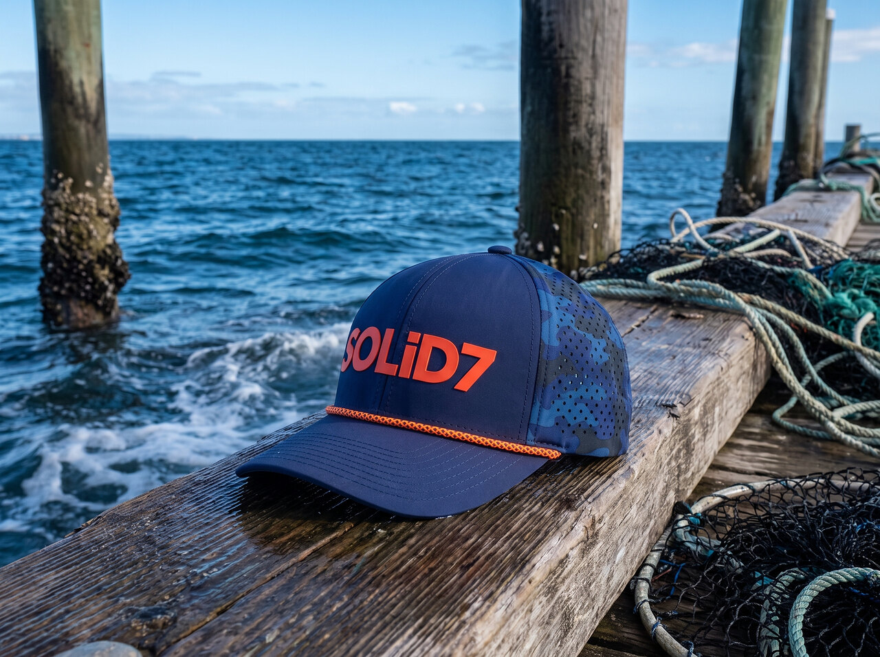 Navy SOLiD7 cap resting on a weathered jetty beam above the sea, fishing rope and nets alongside