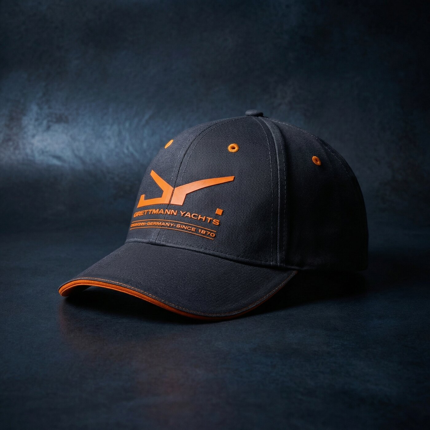 A charcoal Brettmann Yachts cap with an orange mark, under low studio light